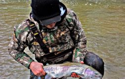 Man in a jacket holding large trout in a stream